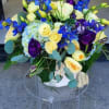 Low arrangement of yellow and cream roses, blue hydrangea, and purple blooms in a white vase on a clear stand