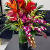 Tall tropical arrangement with purple orchids, red anthuriums and orange flowers in a clear glass vase