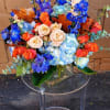 Arrangement of blue hydrangeas, blue delphinium, and orange and peach roses in a clear glass vase