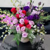 Low bowl arrangement of pink and red roses with purple blooms and orchids in a lush mixed design