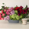 Mixed bouquet of roses, tulips, and anemones in a gray container beside a candle