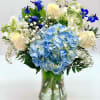 Vase arrangement with blue hydrangeas, cream roses, white blooms, and bright blue flowers
