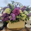 Purple and green mixed flower arrangement in a cream tote-style box on a wooden table