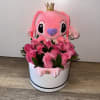 Pink plush character with a crown sitting in a box of pink roses