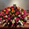 Large multicolored rose and lily casket spray with a purple ribbon bow arranged in an oval shape.