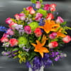Tall mixed arrangement of pink roses, orange lilies, green mums, and blue accents in a white vase with purple ribbon.