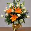 Tall arrangement of orange lilies, white roses, blue flowers, and hydrangeas in a gold vase with a bow