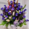 Tall arrangement of purple roses, pink lilies, and mixed blooms in a vase with a sheer ribbon bow