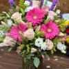 Vase arrangement with hot pink gerbera daisies, pale roses, lilies, and mixed blooms in a green textured glass vase