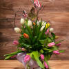 Mixed tulip arrangement with colorful butterfly picks in a clear vase on a wood slice base.
