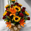 Mixed arrangement of sunflowers, orange lilies, and red roses in a glass vase with a burlap bow