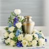 Cremation urn surrounded by white roses, white chrysanthemums, and blue flowers in a low arrangement