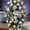 Tall standing spray of white lilies, roses, carnations and blue hydrangeas with bright blue accent blooms.