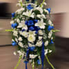 Tall standing spray of blue roses, white lilies, and white mums with blue ribbon on an easel.