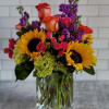 Bright mixed arrangement with sunflowers, roses, and purple blooms in a clear glass vase