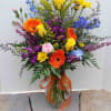 Tall mixed arrangement of bright yellow roses, orange gerberas, and purple and blue flowers in a glass vase
