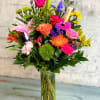 Tall mixed bouquet of bright pink, orange, yellow, and purple flowers in a clear glass vase.