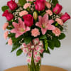 Tall arrangement of hot pink roses, pink lilies, and light pink carnations in a clear vase with a floral bow.