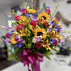 Tall mixed bouquet with sunflowers, pink roses, lilies and purple-blue blooms in a glass vase with magenta bow