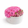 Round white box filled with pink roses and chocolate-covered strawberries drizzled with pink and white.