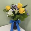 Arrangement of yellow roses, white lilies, and purple asters in a dark vase with a blue ribbon bow
