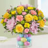 Bouquet of yellow roses, pink chrysanthemums, and carnations in a glass vase filled with pastel eggs.