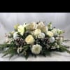 Low white flower centerpiece with pinecones, ornaments, and a gold bow in winter evergreens
