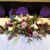 Low centerpiece of pink roses, white blooms, and purple flowers arranged across a banquet table