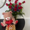 Red rose bouquet in a glass vase with purple blooms, a plush bear with red heart, and a box of chocolates.