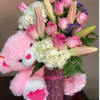 Pink teddy bear hugging a vase of pink roses, lilies, and white hydrangeas