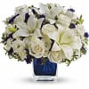 Low arrangement of white lilies and roses with purple accents in a blue cube glass vase