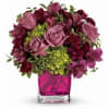 Compact arrangement of mauve roses, dark carnations, and green hydrangea in a bright pink glass cube vase