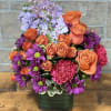 Bright arrangement of orange roses, purple daisies, and pink carnations in a green cube vase with Happy Birthday pick