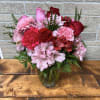 Pink and red roses, carnations, and alstroemeria arranged in a clear glass vase on a wooden table.