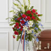 Tall red, white, and blue standing spray with carnations, lilies, and ribbon on an easel beside a casket.