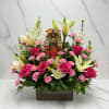 Pink roses and lilies arranged around a religious figurine in a rectangular vase