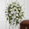 Standing spray of white roses, lilies, and carnations with a white ribbon on an easel beside a wooden casket.