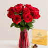 Red roses in a red glass vase with a box of chocolates beside them