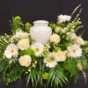 White urn surrounded by cream roses and white daisies in a green floral arrangement