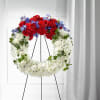 Circular funeral wreath of red, white, and blue flowers on a black stand