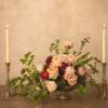 Low floral centerpiece with blush and peach roses on a pedestal, flanked by two lit taper candles