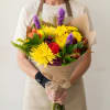Person holding a colorful bouquet of yellow, purple, and red flowers wrapped in brown paper