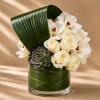 White roses and orchids arranged with a succulent in a glass vase wrapped in green leaves