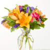 Mixed bouquet of orange lilies, pink roses, and purple filler flowers in a clear glass vase