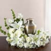 White lilies, roses, and mums arranged in a ring around a silver urn on a dark table.