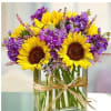 Sunflowers and purple flowers in a clear glass vase with a raffia bow