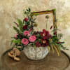 Pink and burgundy floral arrangement in a white ceramic bowl with a gold frame behind it