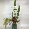 Tall floral arrangement in a teal glass vase with a pink protea and green blooms
