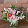 Low glass vase of pink tulips, ranunculus, roses, and white blooms in a rounded bouquet.