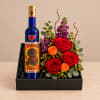 Roses and purple flowers arranged in a black gift box beside a decorated bottle